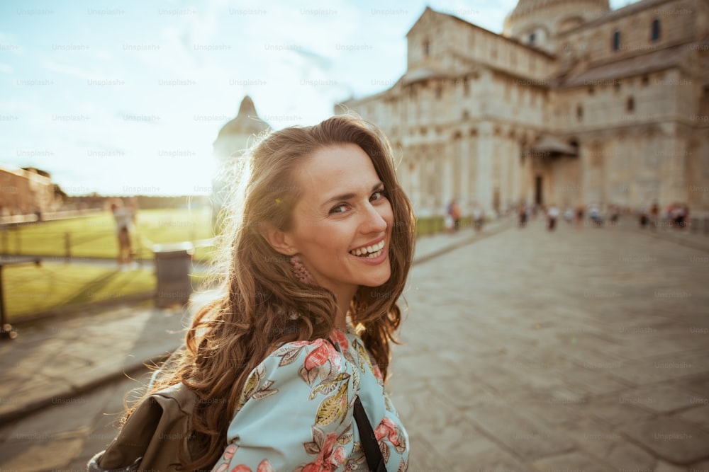 feliz y elegante viajera de mediana edad con vestido floral con mochila que tiene recorrido a pie en piazza dei miracoli en Pisa, Italia.