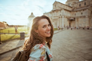 Donna viaggiatrice di mezza età elegante felice in abito floreale con zaino che fa un tour a piedi in piazza dei miracoli a Pisa, Italia.