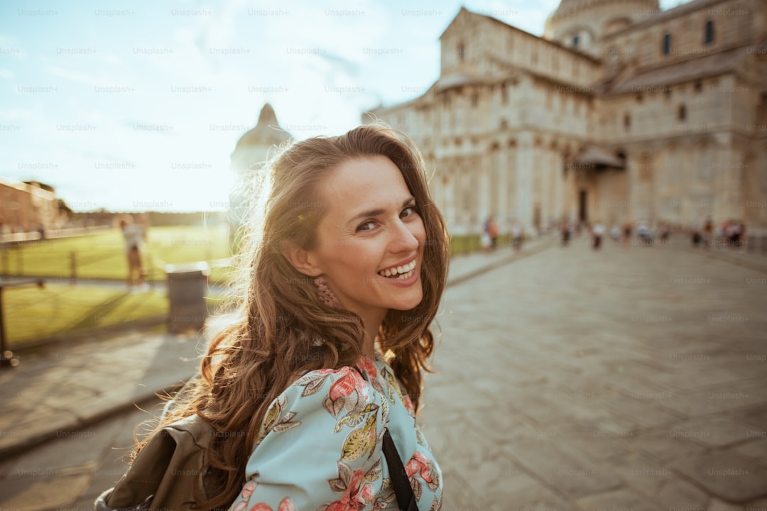 happy elegant middle aged traveller woman in floral dress with backpack having walking tour in piazza dei miracoli in Pi