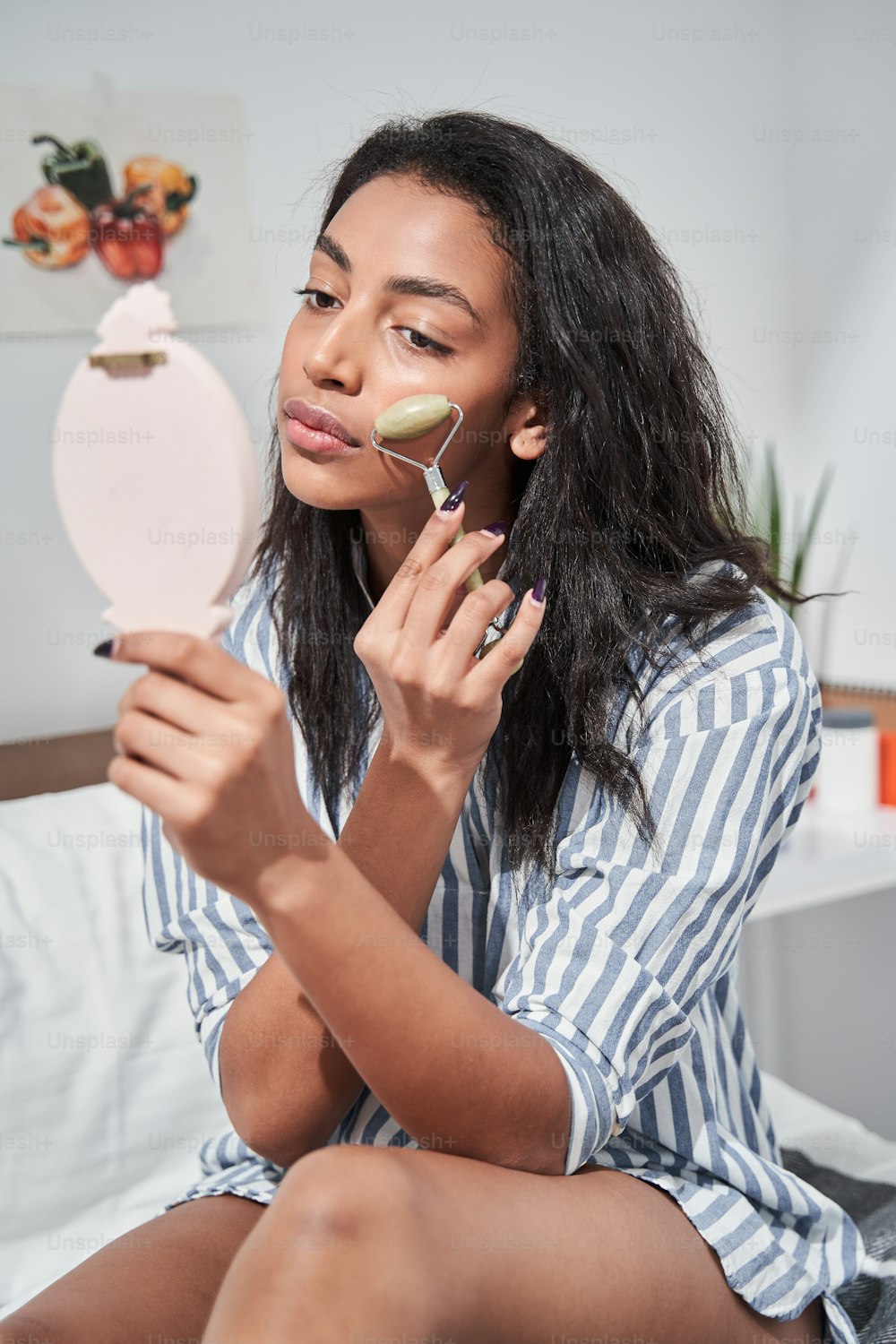 Young brunette woman is holding hand mirror and looking at her reflection with pleasure smile while using jade roller on cheeks at the bedroom. Concept of beauty, self-care, cosmetics