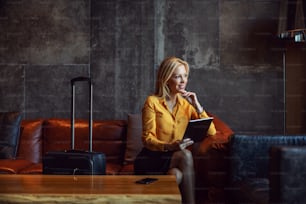 Happy businesswoman sitting in a hall of a hotel and checking in a hotel online. She is on a business trip. Telecommunications, travel, business trip