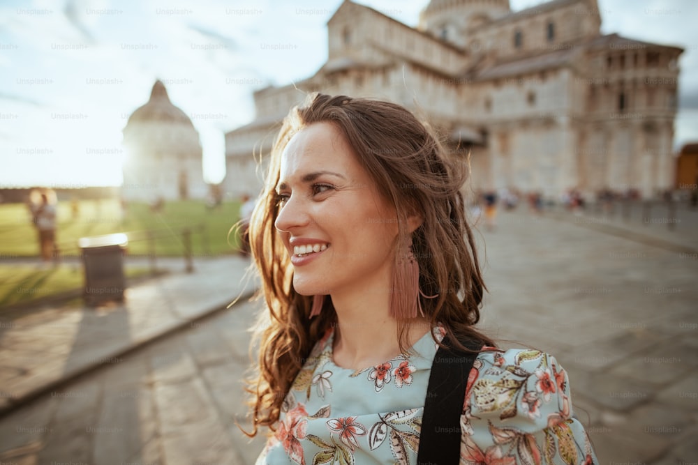 mujer joven feliz en vestido floral con mochila haciendo turismo en la plaza de los milagros en Pisa, Italia.