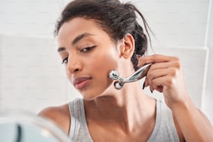 Young woman is holding hand mirror and looking at herself while using jade roller on cheeks at the bathroom. Concept of beauty, self-care, cosmetics