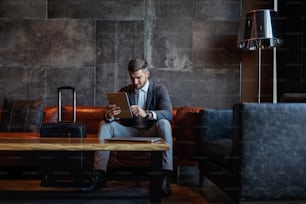 Businessman sitting in a hall of a hotel and using tablet. He is on a business trip.