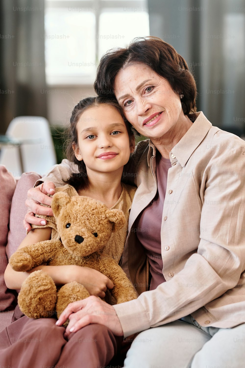 Affectionate mature brunette woman and her cute little granddaughter looking at you with smiles while sitting in front of camera