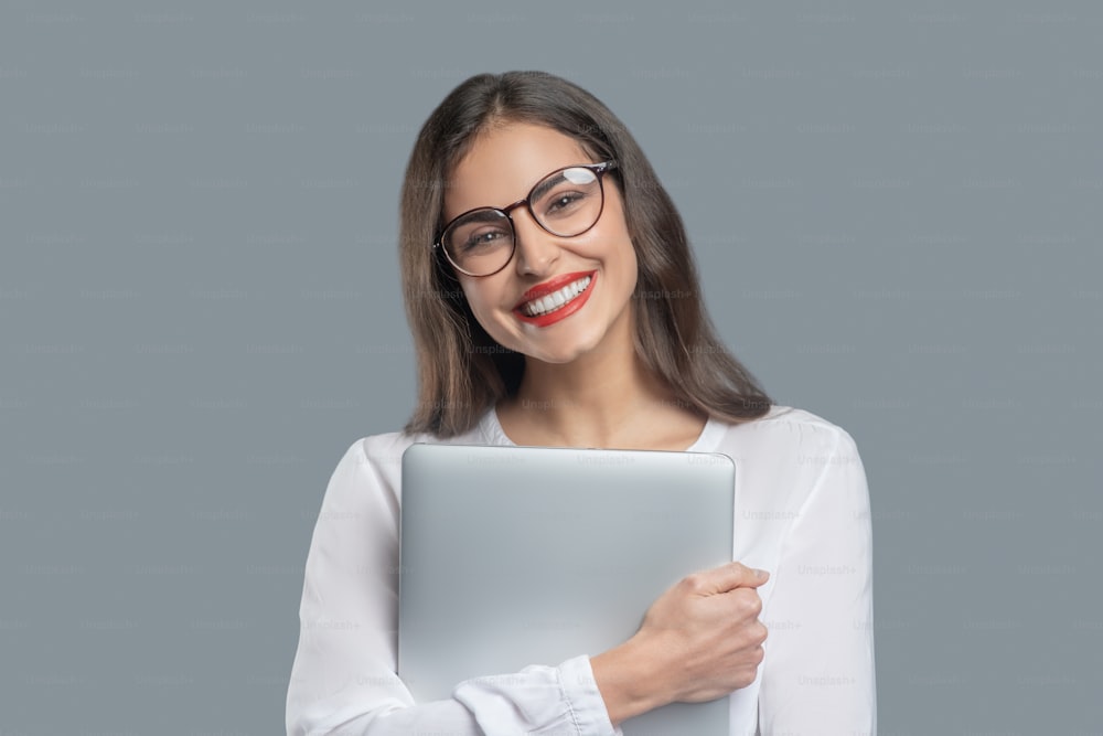 Joy, success. Happy young business woman with white-toothed smile wearing glasses holding laptop tightly to herself