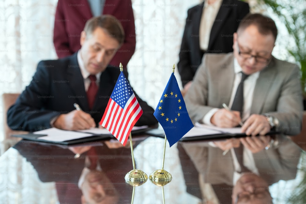 Les drapeaux des États-Unis et de l’Union européenne sur la table contre deux délégués signant un contrat après avoir négocié et conclu un accord