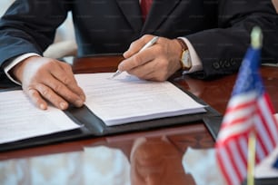 Hände eines reifen amerikanischen Delegierten in formeller Kleidung, der den Vertrag über die Geschäftspartnerschaft unterzeichnet und auf die Unterschrift zeigt, während er am Tisch sitzt