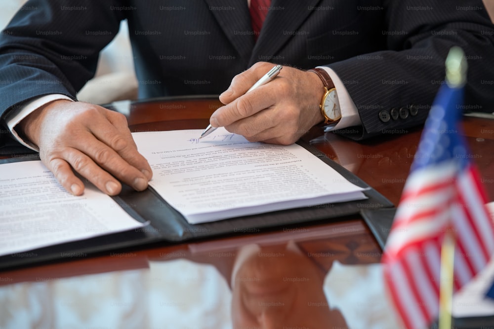 Hands of mature American delegate in formalwear signing contract of business partnership and pointing at signature while sitting by table