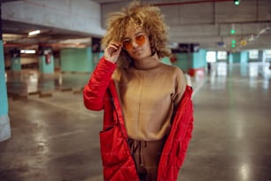 Sérieuse fille hip hop métisse en veste debout dans le garage et ajustant des lunettes de soleil.