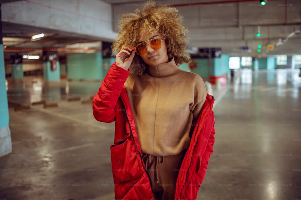 Ernsthaftes gemischtrassiges Hip-Hop-Mädchen in Jacke, das in der Garage steht und die Sonnenbrille anpasst.