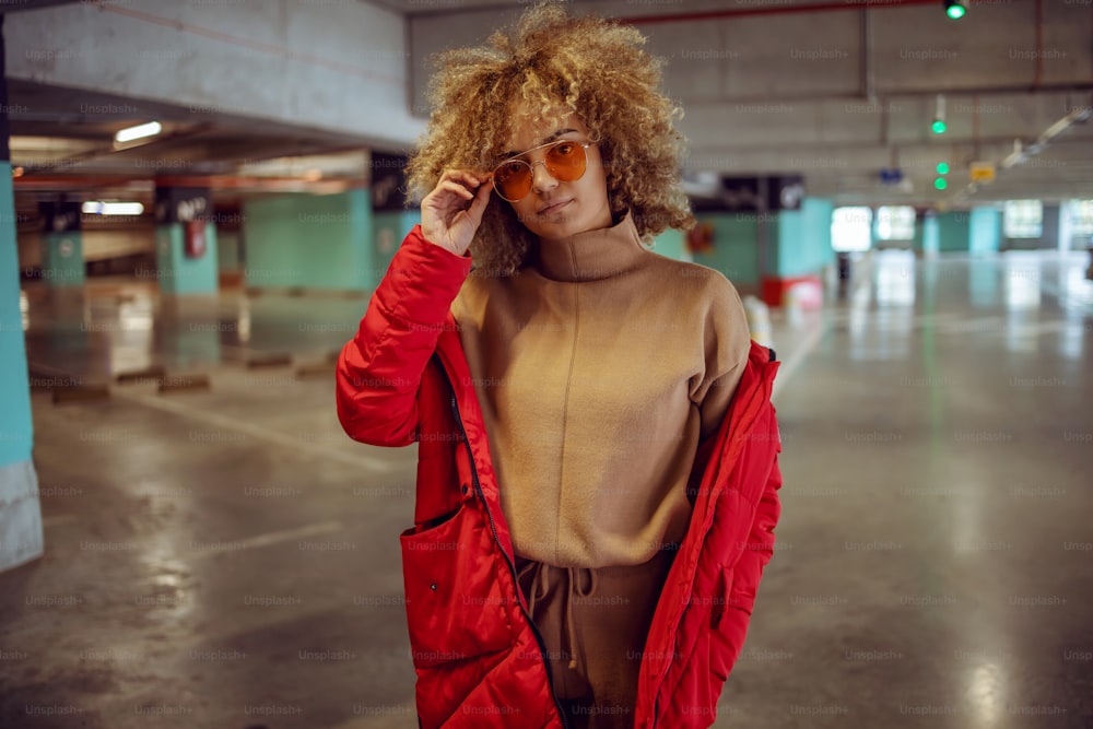Serious mixed race hip hop girl in jacket standing in garage and adjusting sunglasses.