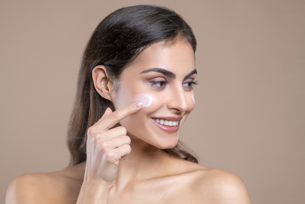 Dab cream. Close-up photo of pretty long-haired woman applying cream to her cheek with her finger in good mood