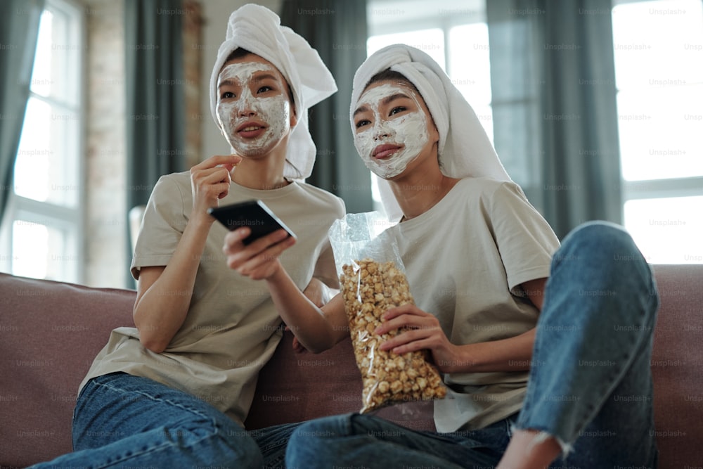 Young happy Asian female twins with towels on heads and clay mask on faces sitting in front of tv set, watching movie and having popcorn