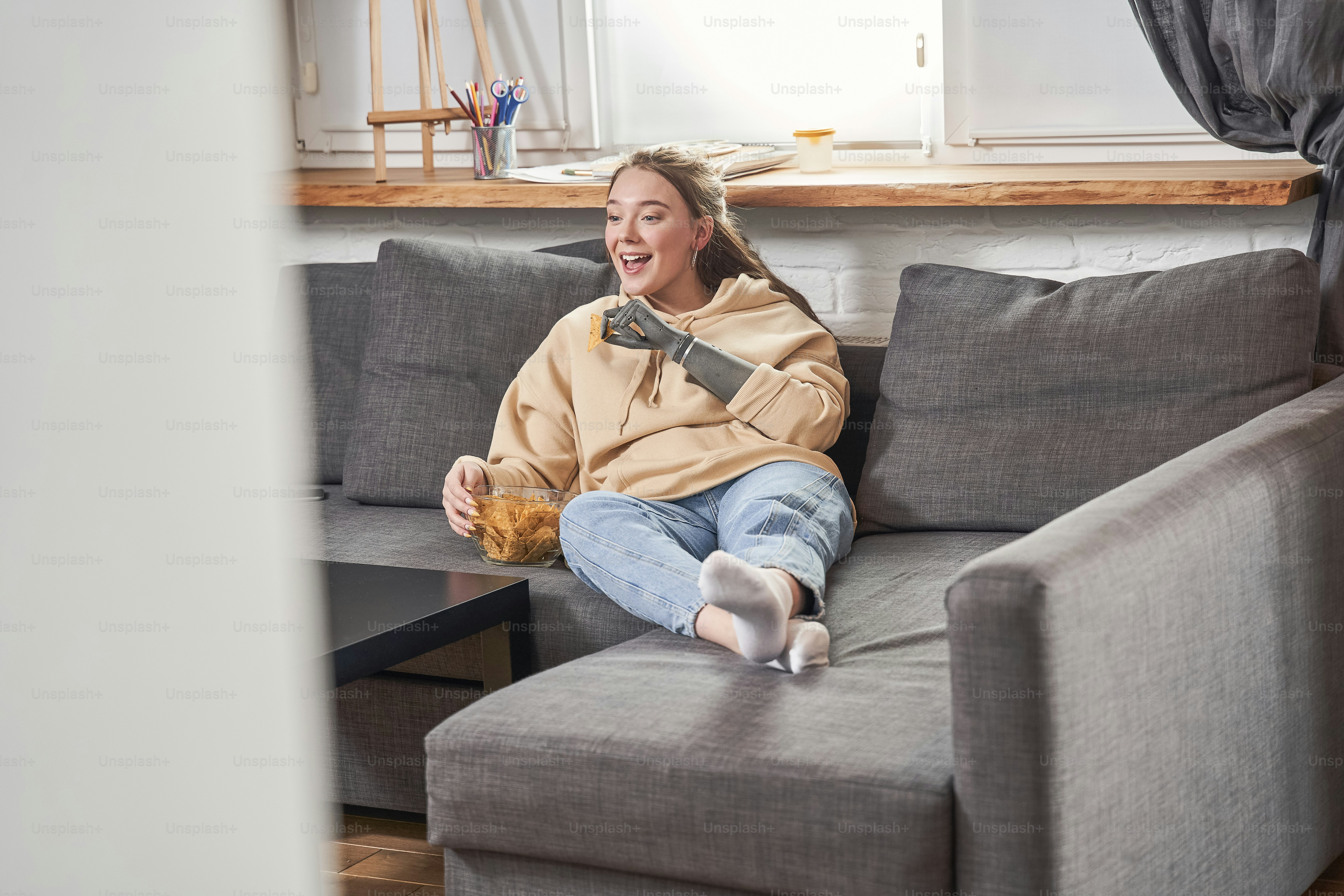 A woman with a prosthetic arm eating chips on a grey sofa