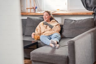 Porträtansicht der kaukasischen fröhlichen Frau mit Implantatarm, die lacht und Pfeffer-Nachos-Chips mit Appetit isst, während sie auf dem Sofa in der Selbstisolation einen Film schaut