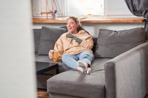 Portrait view of the caucasian cheerful woman with implant arm laughing and eating pepper nachos chips with appetite while watching movie at the sofa at the self isolation