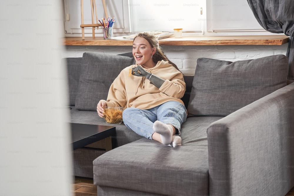 Portrait view of the caucasian cheerful woman with implant arm laughing and eating pepper nachos chips with appetite while watching movie at the sofa at the self isolation