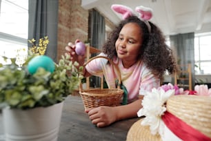 African little girl sitting at the table with basket and playing with Easter egg she celebrating Easter at home