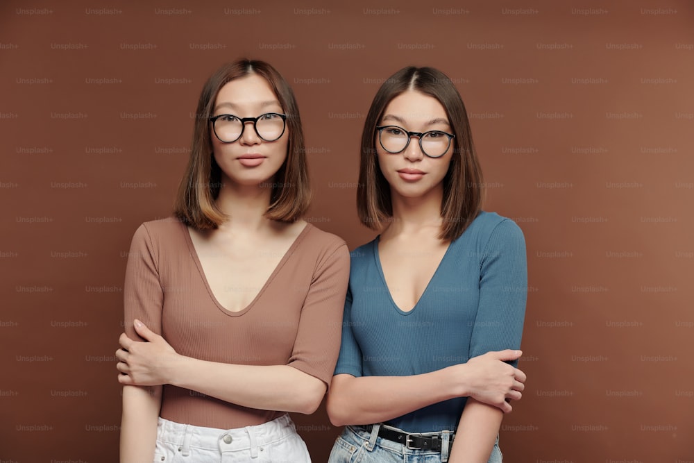 スタジオの茶色の背景にカメラの前で近くに立つプルオーバーと眼鏡をかけた若い真面目な双子の姉妹