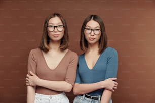 Young serious twin sisters in pullovers and eyeglasses standing close to one another in front of camera against brown background in studio