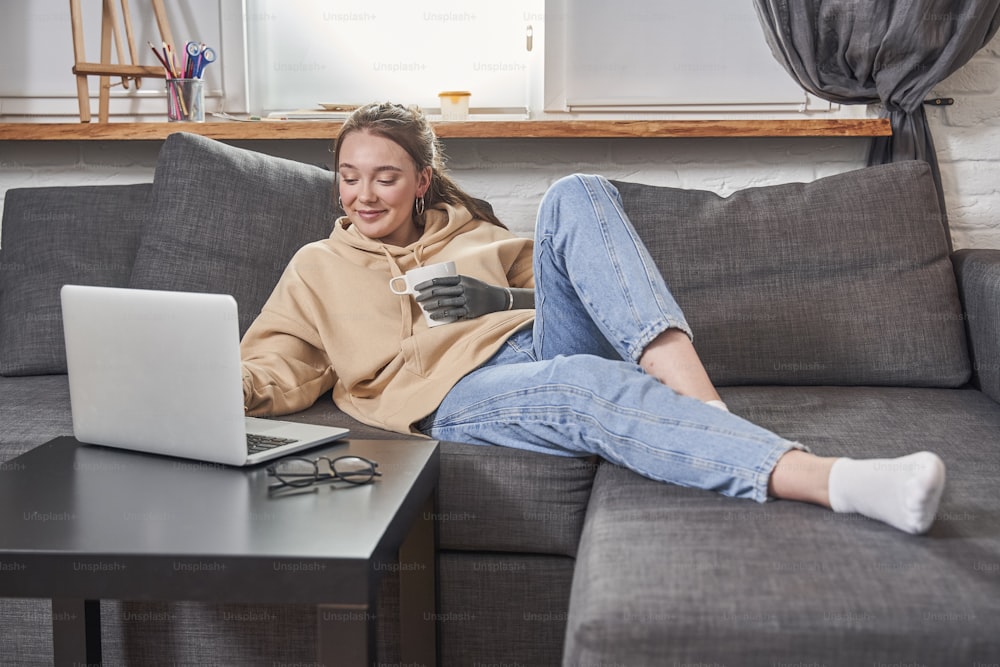 Happy red haired woman with prosthesis arm typing at the laptop keyboard while laying at the sofa and watching movie in living room. Technology and disabled people concept. Stock photo