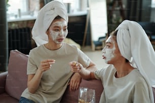 Young Asian female twins with towels on heads and clay mask on faces sitting on couch in front of camera, talking and having popcorn