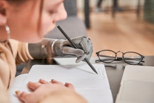 Portrait view of the caucasian teenage girl with prosthesis arm sitting at the table and writing her homework tasks. Woman holding pen at artificial limb. Disabled people concept