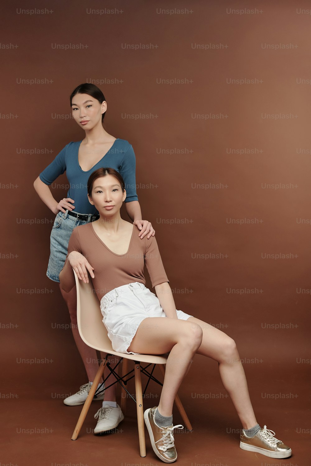 캐주얼웨어를 입은 젊은 진지한 갈색 머리 여성 쌍둥이가 갈색 벽이 있는 스튜디오의 흰색 플라스틱 의자에 앉아 있는 동안 당신을 바라보고 있습니다