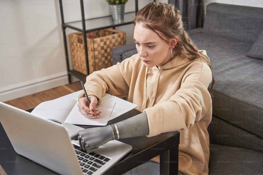 Retrato de la mujer caucásica tranquila con implante de brazo sentada a la mesa con su computadora portátil y escribiendo en el teclado mientras navega por Internet para estudiar. Concepto tecnológico