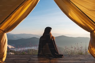 Rückansicht einer Frau, die auf einem Holzbalkon sitzt und einen wunderschönen Blick auf die Berge und die Natur außerhalb des Zeltes beobachtet