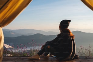 Rear view image of a woman sitting on wooden balcony while watching a beautiful mountains and nature view outside the tent