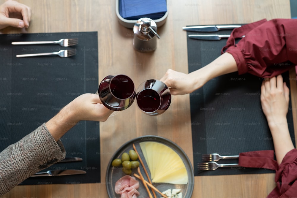 Visão geral das mãos do jovem e as de sua namorada piscando por taças de vinho tinto sobre a mesa servida durante o jantar romântico