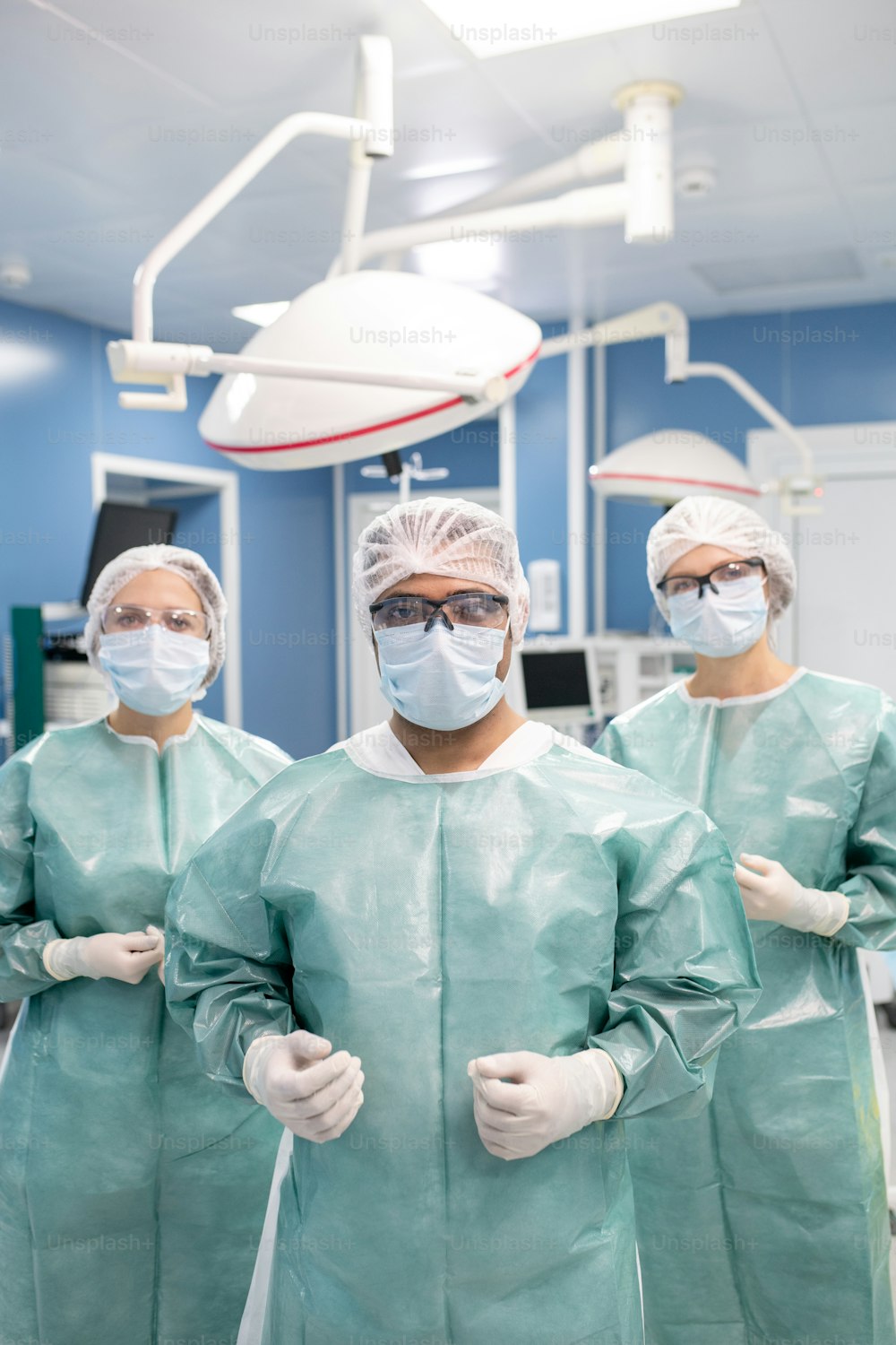Tres cirujanos contemporáneos en ropa de trabajo protectora se preparan para la operación mientras están de pie frente a la cámara en la sala de operaciones