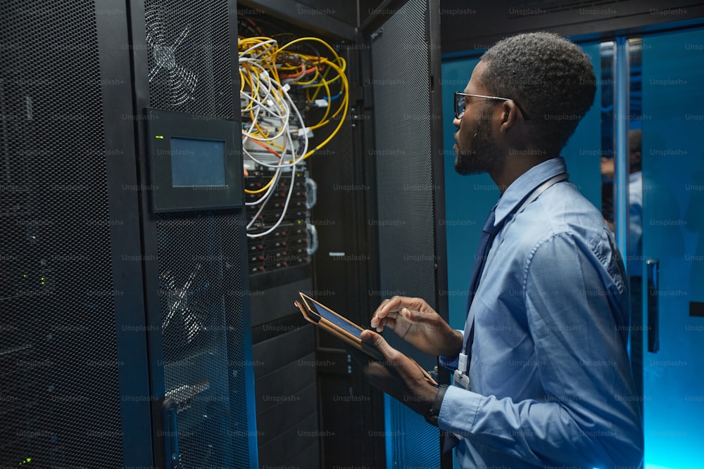 Seitenansichtsporträt eines afroamerikanischen Mannes, der am Serverschrank steht, während er mit einem Supercomputer im Rechenzentrum arbeitet und das Tablet hält, Kopierraum