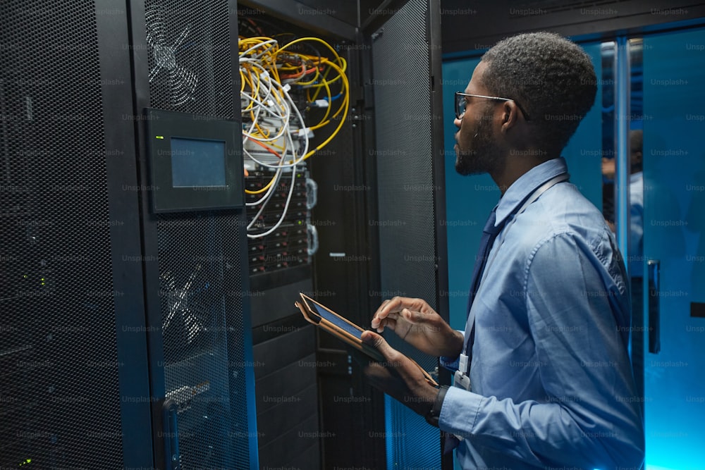 Side view portrait of African American man standing by server cabinet while working with supercomputer in data center and holding tablet, copy space