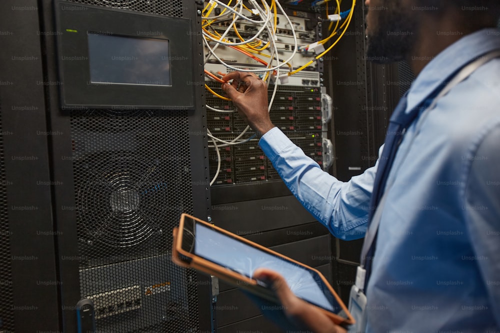 Retrato de recorte de un ingeniero de redes afroamericano que conecta cables en el gabinete del servidor mientras trabaja con una supercomputadora en el centro de datos, espacio de copia