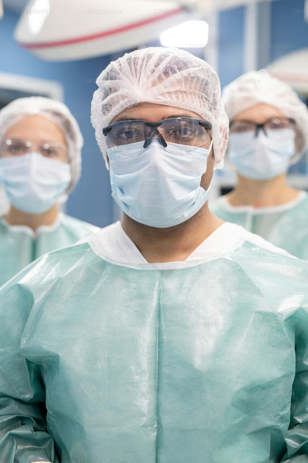 Junger professioneller männlicher Chirurg und seine beiden Assistenten in Schutzuniform, Maske und Brille stehen im Operationssaal