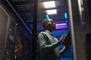 Portrait en contre-plongée d’un ingénieur de données afro-américain travaillant avec un supercalculateur dans une salle de serveurs éclairée par la lumière bleue, espace de copie
