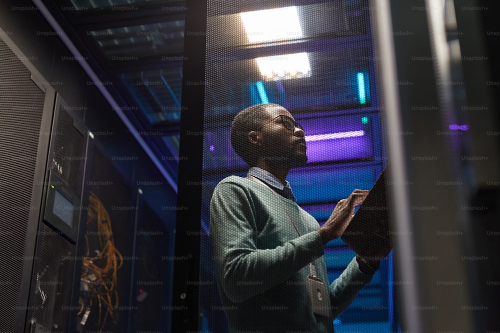 Ritratto a basso angolo di un ingegnere informatico afroamericano che lavora con un supercomputer nella sala server illuminata da luce blu, spazio di copia