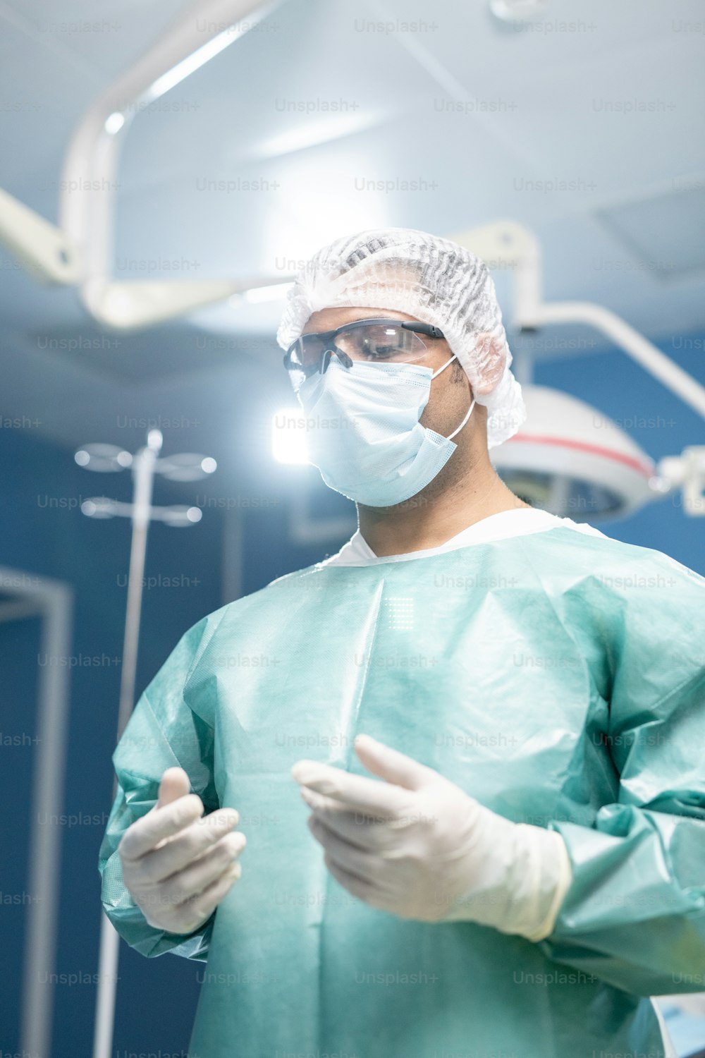 Cirujano profesional masculino con ropa de trabajo protectora, máscara, guantes y anteojos de pie en la sala de operaciones y que va a operar al paciente