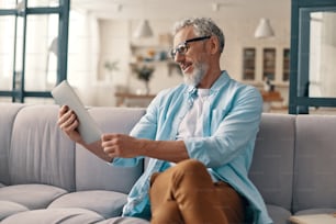 Busy senior man in casual clothing using digital tablet while sitting on the sofa at home