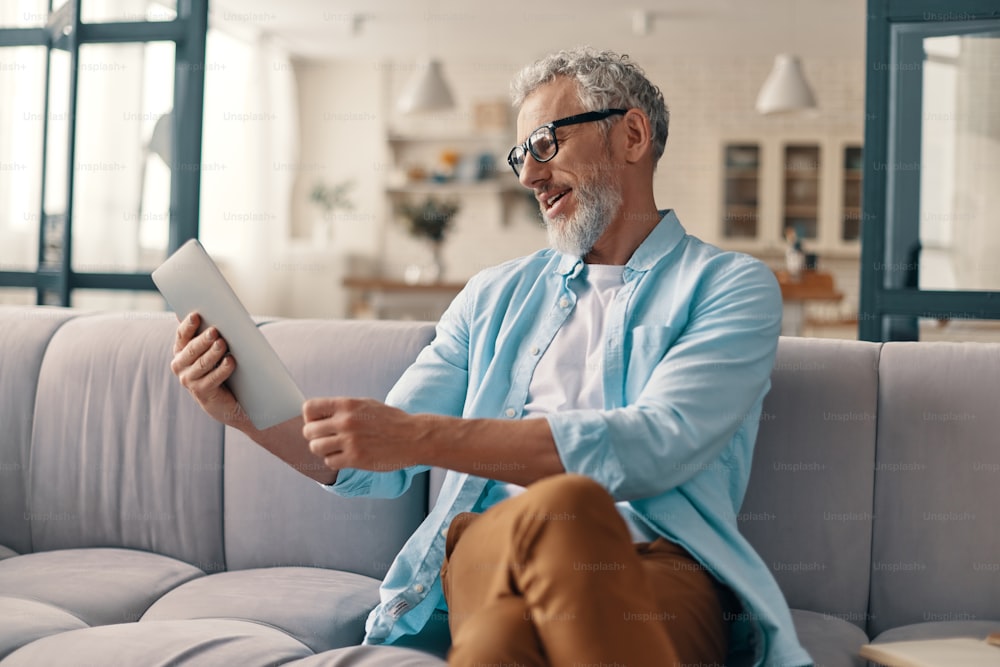 Hombre mayor ocupado con ropa casual usando tableta digital mientras está sentado en el sofá de su casa