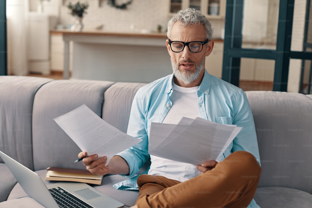 Thoughtful senior man checking the papers with serious face while sitting on the sofa at home