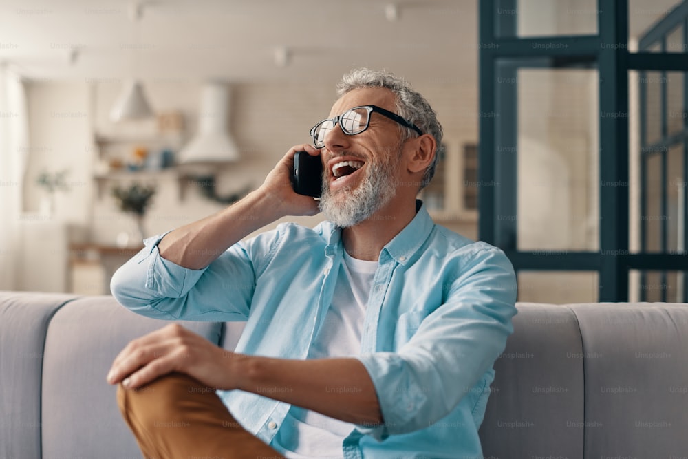 Hombre mayor sonriente con ropa casual y anteojos hablando por el teléfono inteligente mientras está sentado en el sofá de su casa