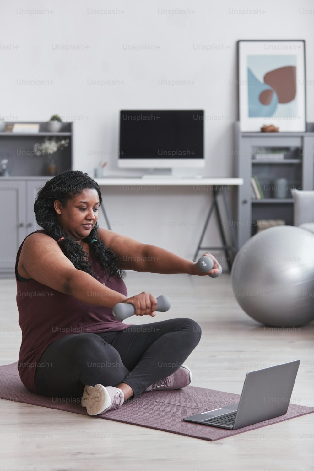 Vertikales Porträt einer kurvigen Afroamerikanerin, die zu Hause mit Hanteln trainiert, während sie auf einer Yogamatte sitzt und Online-Trainingsvideos ansieht.