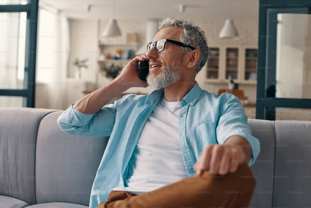 Hombre mayor relajado con ropa informal y anteojos hablando por el teléfono inteligente mientras está sentado en el sofá de su casa