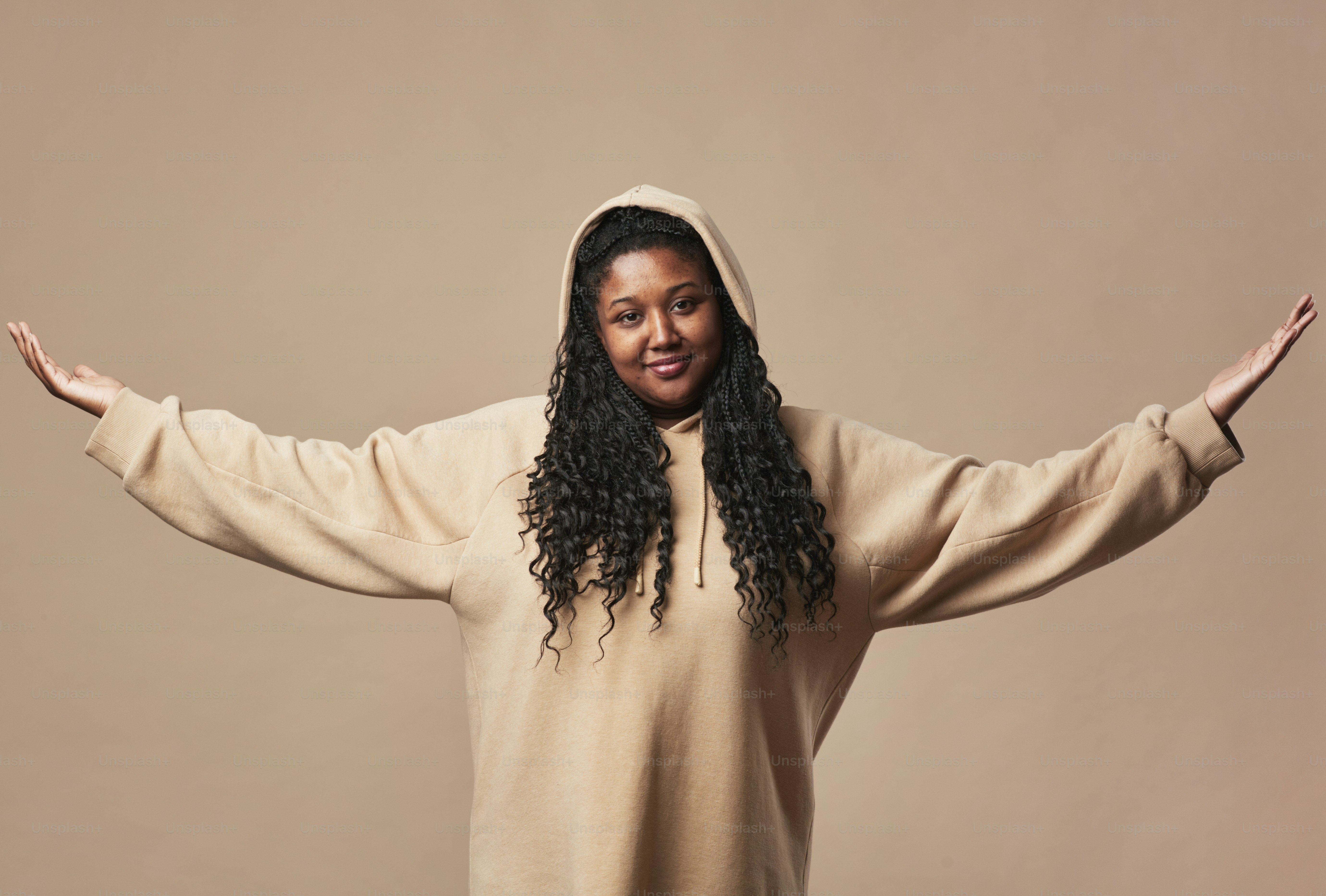 Seorang wanita dengan kepang panjang gelap mengenakan hoodie krem dengan tangan terentang