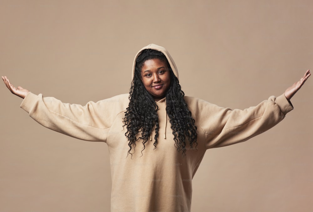 Minimal waist up portrait of confident curvy woman looking at camera and wearing comfortable hoodie while standing with arms raised against neutral beige background in studio, copy space