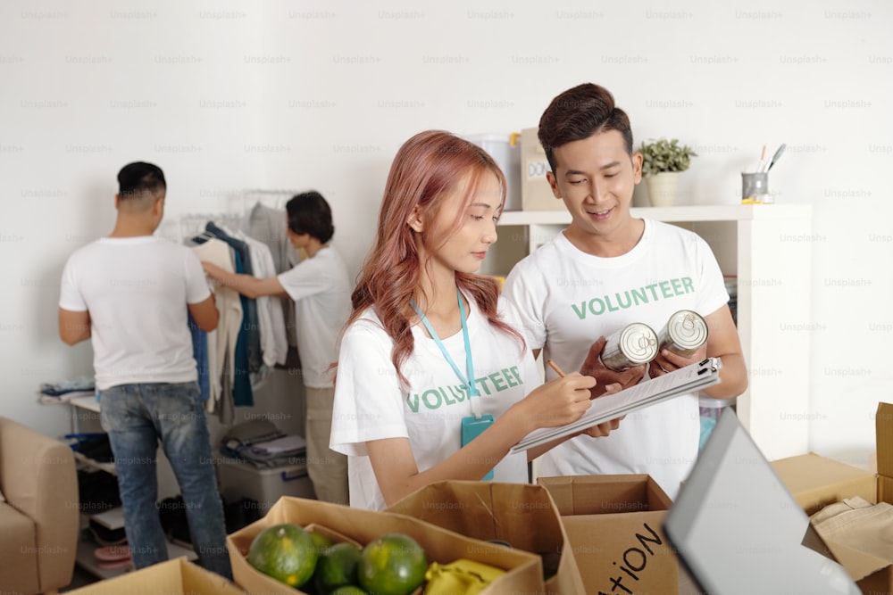 Volunteers working in charitable foundation taking notes in document and describing all food and clothes they are packing for people in need